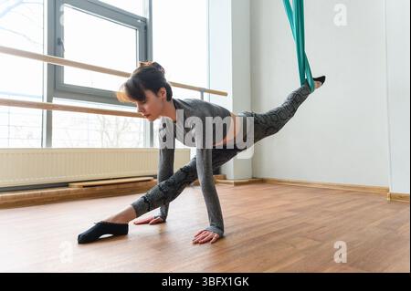 Woman in gray active wear performs a low split stretch on the floor, with one leg elevated and supported by teal aerial silk in a bright studio with w Stock Photo