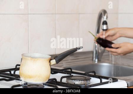 Turk on the stove and a woman washing an eggplant under the tap in the kitchen, cooking, household environment, making coffee Stock Photo