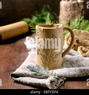 Decorating ceramics. Raw clay mug with a branch imprint. Children's creativity. Amateur ceramics. Rough fabric, tools, branches on wooden table. Eco d Stock Photo