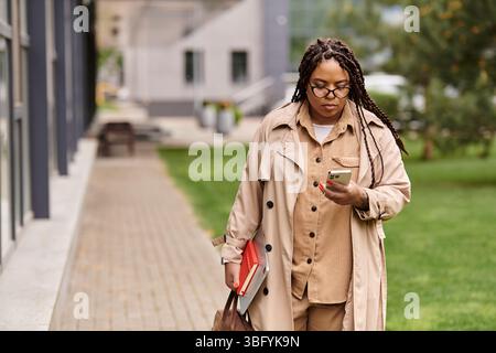 A university teacher navigates campus pathways dressed in a stylish trench coat, checking her phone. Stock Photo