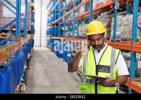 Man wearing yellow helmet and vest using clipboard speaking on phone in warehouse aisle, copy space Stock Photo