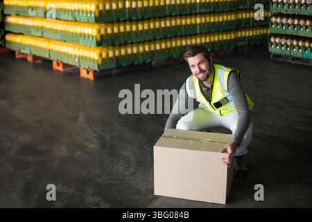 Male warehouse worker wearing vest kneeling in warehouse lifting box beside crates, copy space Stock Photo