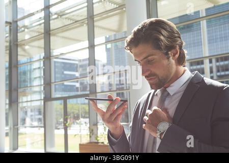 Man speaking into smartphone while wearing dark suit near glass walls in office lobby, copy space Stock Photo