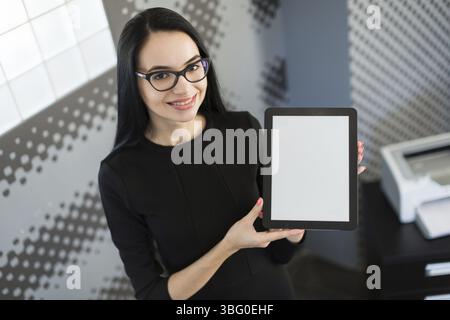 Office on background, beautiful young brunette caucasian businesswoman in black dress and glasses hold red paper folder, show empty tablet, smiling Stock Photo