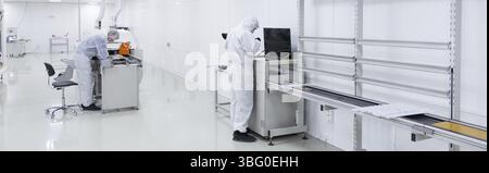 Two factory workers in white lab suits, shoe covers and black latex gloves, working with some modern equipment in a very clean room. panoramic picture Stock Photo