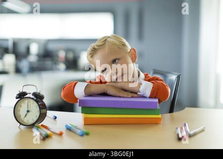 Cute blonde caucasian kid sit at the table with colorful folders and alarm clock and relies on the colorful folders, bored Blurred office background, Stock Photo
