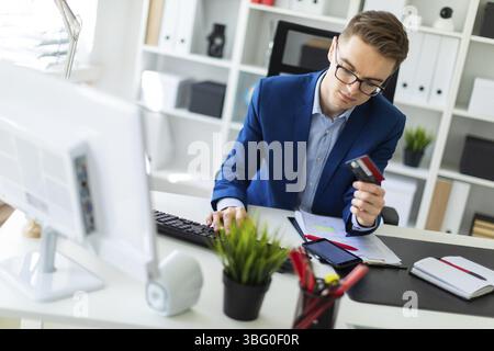 A young man is sitting at a table in the office, holding a bank card in his hand and typing on a computer Stock Photo