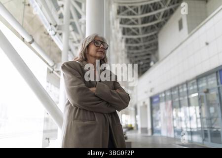 A confident middle-aged businesswoman stands with arms crossed in a sophisticated office environment, reflecting on her professional experiences. The Stock Photo