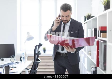 The man is standing near the rack in the office, talking on the phone and holding a folder in his hand Stock Photo