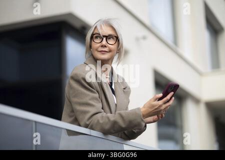 A senior businesswoman stands outside a contemporary building, holding her smartphone. She appears thoughtful and engaged, soaking in the sunlight Stock Photo