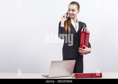 Attractive young caucasian businesswoman with ponytail in black suit stand near the office desk with red paper folders in hands and talk a phone, smil Stock Photo
