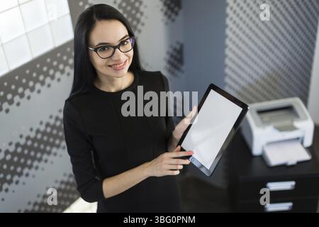 Office on background, beautiful young brunette caucasian businesswoman in black dress and glasses hold red paper folder, show empty tablet Stock Photo