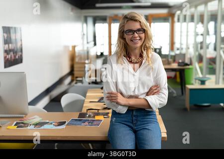 Female professional sitting on desk edge in studio office wearing white blouse with printed photos Stock Photo