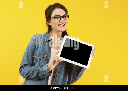 Portrait of attractive brunette young woman in denim shirt and glasses isolated on yellow background with copyspace presenting white tablet pc adverti Stock Photo
