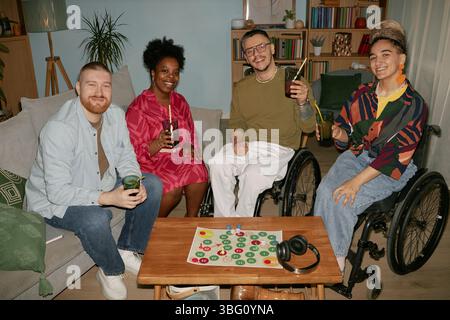 Diverse group of young adults and middle aged people with disability sitting together celebrating birthday, smiling and holding drinks around board game in living room Stock Photo