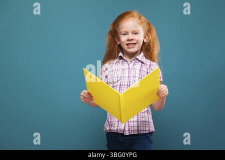 Isolated on blue, attractive caucasian redhead child in white plaid shirt and blue jeans, with long rusty hair, hold yellow paper folder, opened folde Stock Photo
