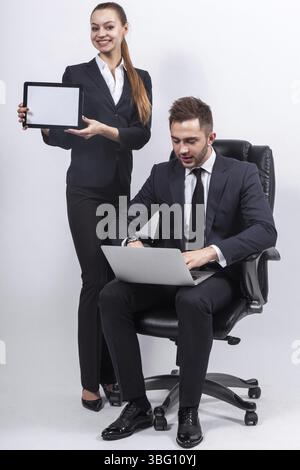 Couple of attractive young caucasian businesswoman with ponytail in black suit with empty tablet in hand and businessman in black suit with laptop Iso Stock Photo