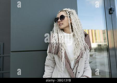 A woman with white dreadlocks is leaning against a wall in an urban setting. She appears relaxed as she gazes into the distance, immersed in her thoug Stock Photo