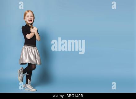 Isolated on blue, red-haired girl in black sweater with zipper, black tights, silver skirt and sneakers, holding her hands and braids crossed. open mo Stock Photo