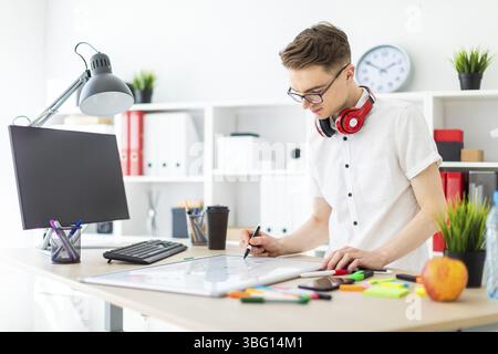 A young man in glasses stands near a computer desk. A young man draws a marker on a magnetic board. On the neck, the guy's headphones hang Stock Photo