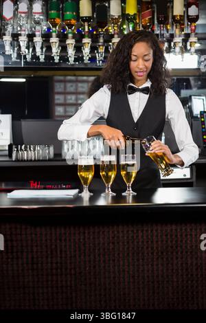 African American woman wearing vest and bowtie pouring beer from bottle into glasses at bar counter Stock Photo