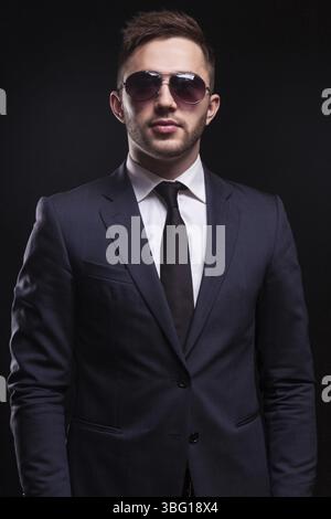 Young attractive brunette caucasian businessman in black suit, costly watch, big sunglasses and tie stand Isolated on black, vertical picture Stock Photo