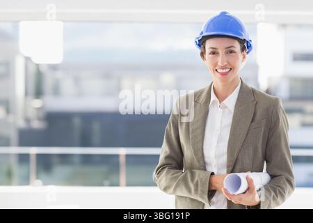 Woman holding plans wearing blazer, blue safety helmet on balcony with glass railing, copy space Stock Photo