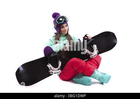 Isolated on white, brunette pretty young caucasian woman in purple ski costume, blue snow boots and blue ski glasses siting cross-legged with black sn Stock Photo