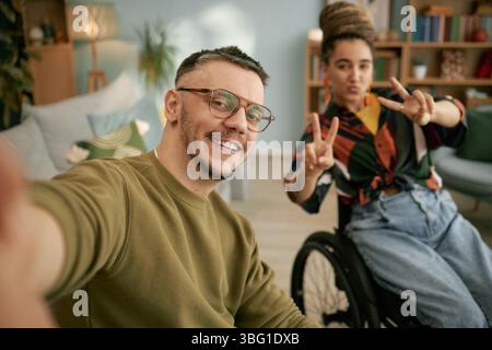 Portrait of cheerful adult man taking selfie while female friend using wheelchair making peace signs during birthday celebration at home Stock Photo