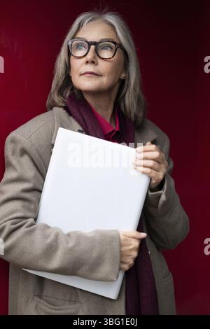 An elderly woman with stylish glasses and gray hair stands with a confident posture, holding a folder close to her chest against a vibrant red backgro Stock Photo