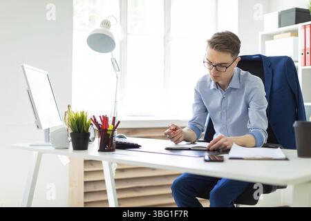 A handsome young man with glasses, blue trousers and a light shirt is working in the office. On the back of the chair hangs his jacket. photo with dep Stock Photo