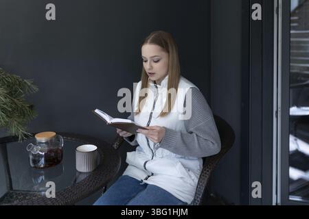 A young woman engages in reading a book at a small table while sipping coffee in a modern indoor setting. The winter light creates a warm atmosphere, Stock Photo