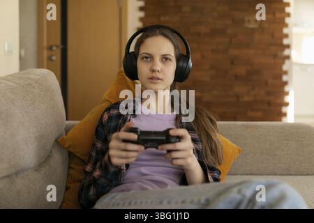 A young woman is deeply focused on her gaming session indoors. She wears headphones while comfortably sitting on a couch with a controller in hand, su Stock Photo