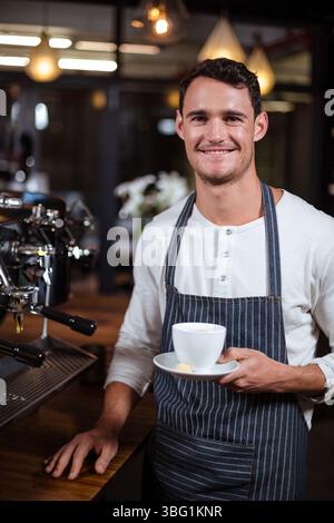 Barista is standing behind counter beside espresso machine holding cup on saucer with sugar cube Stock Photo