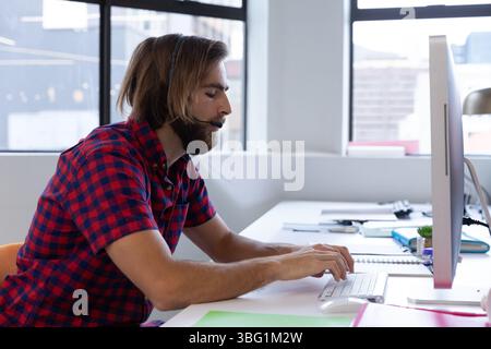 Bearded man wearing checkered shirt typing on keyboard at office desk with headset and monitor Stock Photo