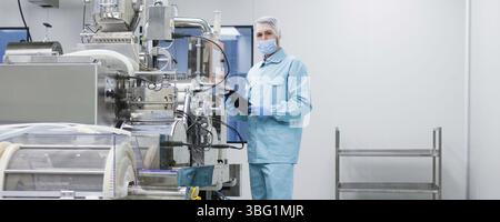 Widescreen picture, caucasian scientist in blue lab uniform configure manufacture machine with shafts and control panel, checking readings, look at ca Stock Photo