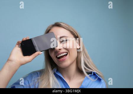 Close-up of a happy european blonde girl holding a smartphone screen with a mockup for an application near her face on a blue background Stock Photo