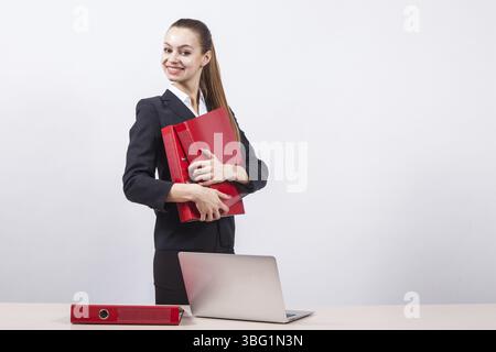 Stylish young business woman designer sites in a business suit stands next to a table with a laptop on a white background and holding a red folder Stock Photo