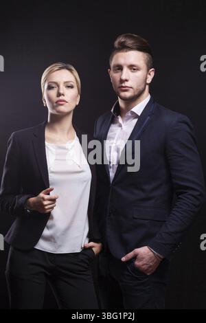 Couple of young serious brunette caucasian lady in grey shirt and businessman in black suit, costly watch and tie stand, look at camera Isolated on bl Stock Photo