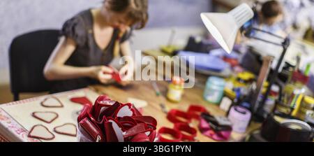 Brown-haired woman manufacturing heart-shaped red purses under the lamp. purses are in focus. blurred background Stock Photo