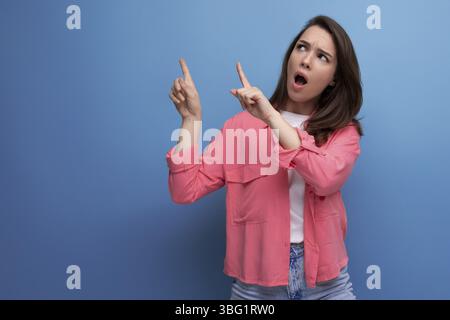 Shocked black haired lady in casual style pointing finger at wall in studio background Stock Photo