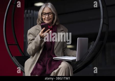 A senior businesswoman engages in work on her laptop outside, occasionally checking her smartphone. She is dressed in professional attire and appears Stock Photo