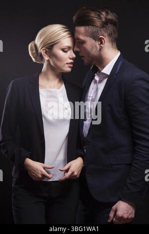 Couple of young serious brunette caucasian lady in grey shirt and businessman in black suit, costly watch and tie stand, close contact Isolated on bla Stock Photo