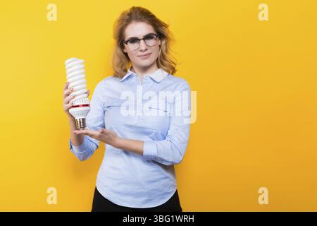 Portrait of attractive blonde woman in blue shirt and eyeglasses holding spiral light bulb isolated on yellow background advertising energy saving con Stock Photo