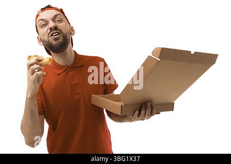 Charismatic courier with a box of pizza in an orange t-shirt and baseball cap on a white background eats a pizza from an order Stock Photo