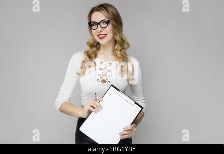 Portrait of attractive curly-haired businesswoman in white top and eyeglasses isolated on grey backgroung holding blank paper list clipped to pad and Stock Photo