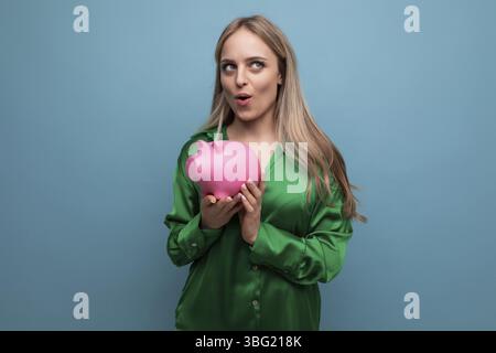 Europaeisch ueberrascht stilvolle blonde maedchen mit piggy bank auf blauem hintergrund Stock Photo