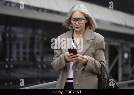 A senior businesswoman dressed in a smart outfit walks outdoors, focused on her smartphone. The background features modern architecture, highlighting Stock Photo
