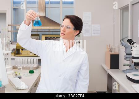 Female scientist holding up conical flask in lab inspecting blue solution wearing goggles Stock Photo