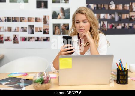 Woman sitting at wooden desk in creative studio using laptop with color wheel, sticky notes nearby Stock Photo
